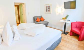 A modern hotel room with a comfortable bed and a cozy seating area. The decoration is bright and inviting, with a small table and a television.