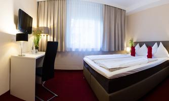 A cozy hotel room with a double bed and two red pillows. The room features a desk and large windows that provide plenty of light.