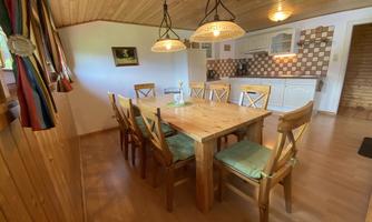A bright dining room with a large wooden table and chairs. The wall has wooden paneling and the kitchen is well equipped.