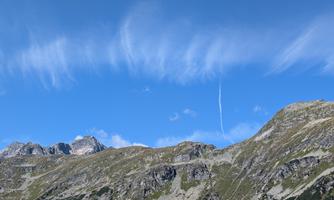 A picturesque mountain panorama under a clear blue sky. The gentle hills and majestic peaks are surrounded by light clouds.