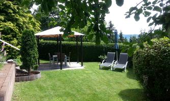 A beautiful garden with a sun terrace and loungers. The lawn is green and surrounded by trees.