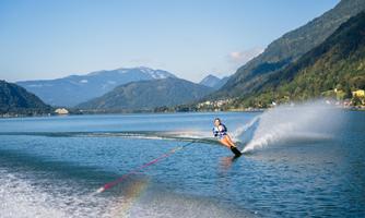 A person is water skiing on a clear lake. In the background, green mountains and a blue sky can be seen.