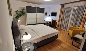 A modern bedroom with a large bed and wooden flooring. The furniture is kept simple, and there is a small workspace as well as a cozy armchair.