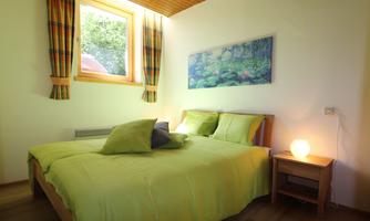A cozy bedroom with a double bed and green bedding. A window with curtains and a wooden desk complete the room.