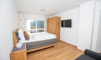 A modern bedroom with a large bed and a TV on the wall. There is bright wood and a window with a view outside.