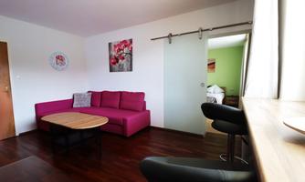 A modern living room with a pink sofa and a round table. In the background, another room is visible, painted in a green tone.