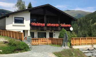 A cozy house with a balcony where colorful flowers bloom. The surrounding landscape is green and mountainous.