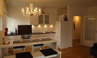 A modern kitchen with an elegant chandelier and a large dining area. The decor is bright and inviting, with wooden flooring and white furniture.