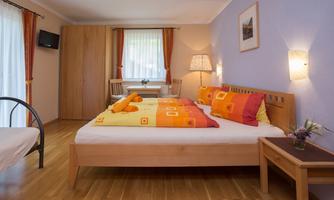 A cozy bedroom with a large bed and colorful pillows. There is a table and a wardrobe, as well as bright curtains and warm wooden flooring.