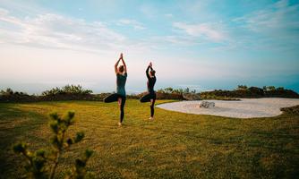 Two women are practicing yoga outdoors on a green meadow. The sky is clear and there is a calm, peaceful atmosphere.