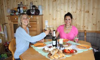 Two women toast with glasses at a table laid with various foods. In the background, a wooden-paneled wall is visible.