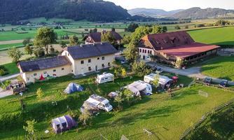 An idyllic camping landscape with several motorhomes and tents. In the background, there is a large farmhouse and gentle hills.