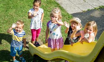 Five children are happily waving from a slide. They are standing on a meadow and enjoying a sunny day outdoors.