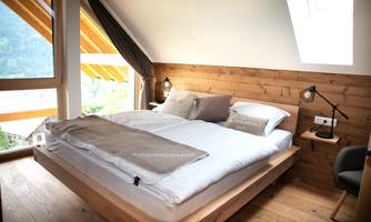 A cozy bedroom with a large double bed and wooden details. Large windows let in plenty of daylight and offer a view of nature.