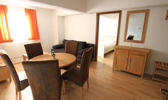 A bright living space with a round table and chairs. In the background, a sofa and a bedroom are visible.