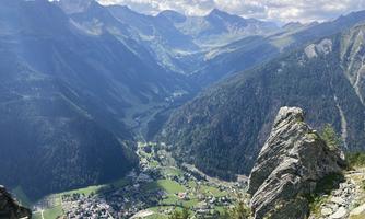 An impressive mountain landscape with green valleys and high peaks. In the foreground, a rock is visible, while the village is visible in the distance.