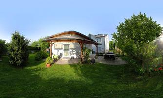 A cozy garden with a wooden pavilion and vibrant plants. In the background stands a modern house under a clear sky.
