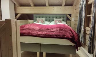 A cozy bedroom with a large bed and a red blanket. The walls are clad in wood and there are windows with curtains.