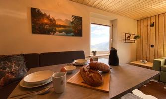 A cozy dining area with a wooden chair and a table that has fresh bread, dishes, and a drink on it. A picture of a picturesque landscape is hanging on the wall.