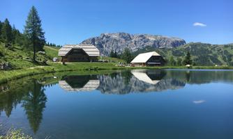 A calm lake surrounded by mountains and greenery. Two traditional huts reflect in the clear water.