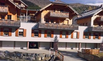 A modern building in alpine style with wooden cladding and balconies. The surroundings are characterized by mountains and natural elements.