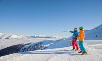 Two skiers stand on a snow-covered slope and look into the distance. In the background, majestic mountains and a clear blue sky can be seen.