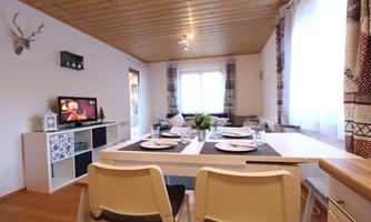 A cozy living room with a dining table and modern chairs. The space is designed in a friendly manner and has large windows that let in plenty of light.