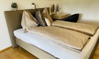 A cozy bedroom with a large bed and beige colored bedding. There are some plants on the nightstand and a modern chair.