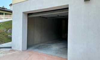 An empty garage with gray walls and an asphalt floor. The entrance is open and leads into a dark room.