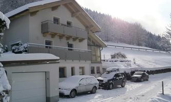 A large house in winter, surrounded by snow. The road is covered in snow and several cars can be seen.