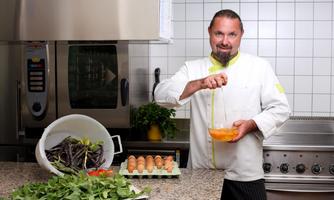 A chef in a modern kitchen is preparing fresh ingredients. He is cracking eggs into a bowl, and around him are arranged vegetables and eggs.