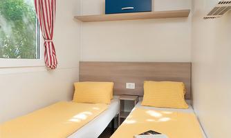 A simple room with two single beds and yellow bedcovers. There is a window with red curtains and an open shelf above the bed.