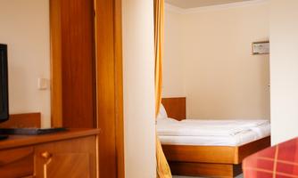 A cozy room with wooden furniture and a bed in the background. The walls are light, and there is a table as well as a television in the room.