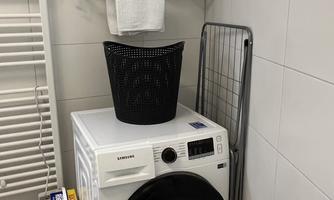 A laundry room with a washing machine and a laundry basket. On a shelf, there are folded towels.