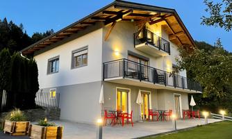 A modern house with a large balcony and a terrace. Red chairs and umbrellas are set up outdoors, surrounded by a well-maintained lawn.