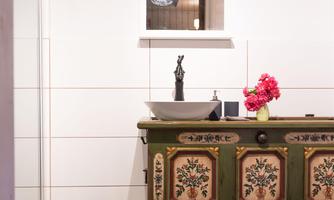 A stylish bathroom with a colorful tiled floor and an old, green cupboard. On the cupboard sits a wash basin and a vase with red flowers.