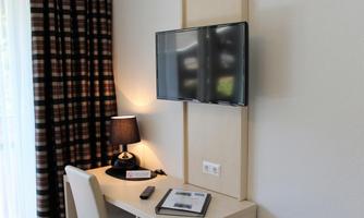 A cozy workspace with a desk, a television on the wall, and a lamp. The curtains are designed in a modern pattern.