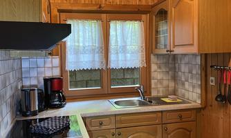 A cozy kitchen with wooden paneling and windows decorated with curtains. The furnishings include a stove, a sink, and a coffee machine.