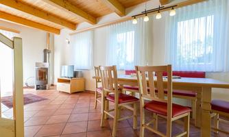 A bright dining room with wooden ceilings and large windows. A table with chairs, a sofa, and a fireplace create a cozy atmosphere.