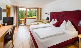 A modern hotel room with a large bed and a cozy seating area. Through the window, a green landscape with trees is visible.