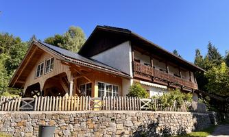 A beautiful house with wooden cladding and a garden. It is situated by a stone wall and is surrounded by trees.