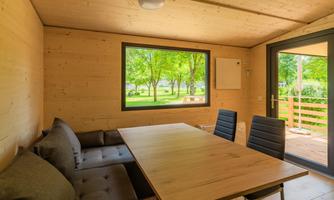 A modern, bright room made of wood with a large table and black chairs. Through the window, one looks out onto a green outdoor area with trees.