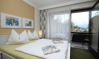 A cozy bedroom with a double bed and colorful cushions. The large windows offer a view of the balcony and the surrounding landscape.