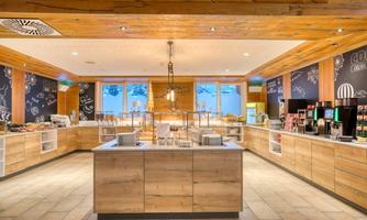 A modern restaurant with wooden decor and an open buffet counter. Large windows provide a view outside and a lot of natural light.