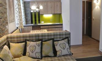 A modern living room with a corner featuring a plaid sofa and decorative cushions. The open kitchen has wood-colored cabinets and a green wall.