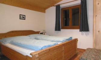 A cozy bedroom with a wooden bed and two pillows. The room has windows with curtains and a wooden ceiling.