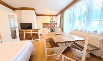 A bright and modern apartment with a kitchenette and a wooden dining table. Large windows provide plenty of daylight.