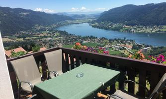 A cozy terrace with a green table and chairs offers a view of a clear lake surrounded by mountains. Colorful flowers beautify the surroundings and create an inviting atmosphere.