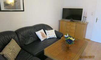 A cozy living room with a black leather sofa and a wooden table. A television and a flower vase are visible in the room.