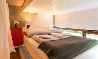 A modern bedroom with a cozy bed and minimalist design. The large windows provide plenty of daylight and a view of nature.